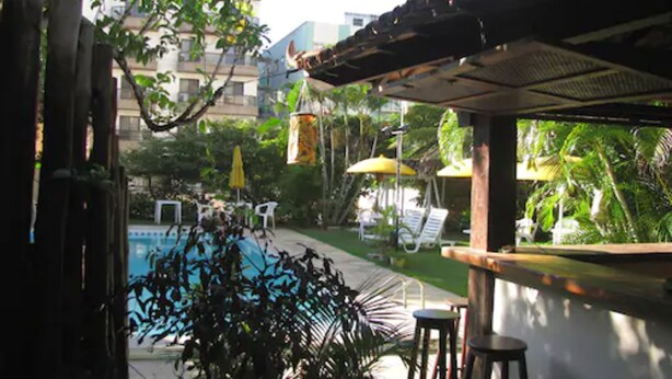 A poolside area with lounge chairs and umbrellas, surrounded by greenery and a building in the background. - Hotel La Brise (Cabo Frio)