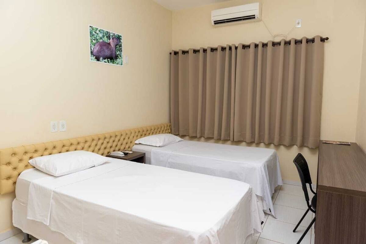 Habitación de hotel con dos camas individuales, un escritorio pequeño, una silla y una ventana con cortinas.