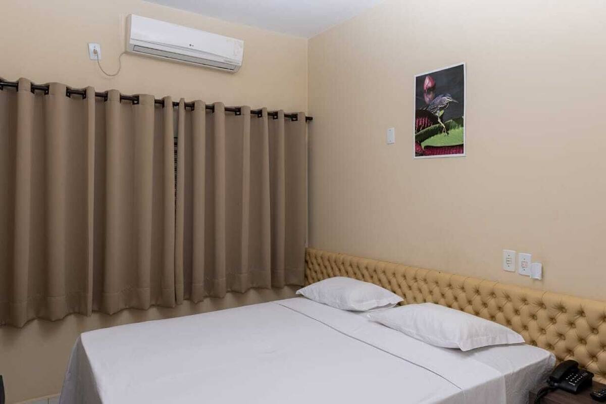 Habitación de hotel con cama, cortinas color beige, aire acondicionado de pared, un cuadro enmarco y un teléfono.