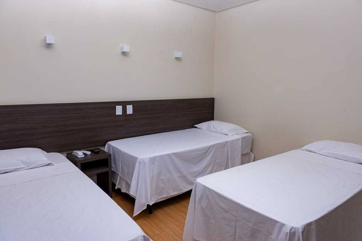 Habitación de hotel con dos camas individuales, cabecera de madera, mesita de noche con teléfono y tres interruptores de luz en la pared.
