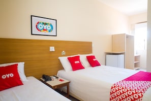 Habitación de hotel con dos camas, cada una con un almohadón rojo que muestra el logotipo del hotel, cabecero de madera, mesita de noche pequeña con teléfono y control remoto, y un cuadro en la pared.