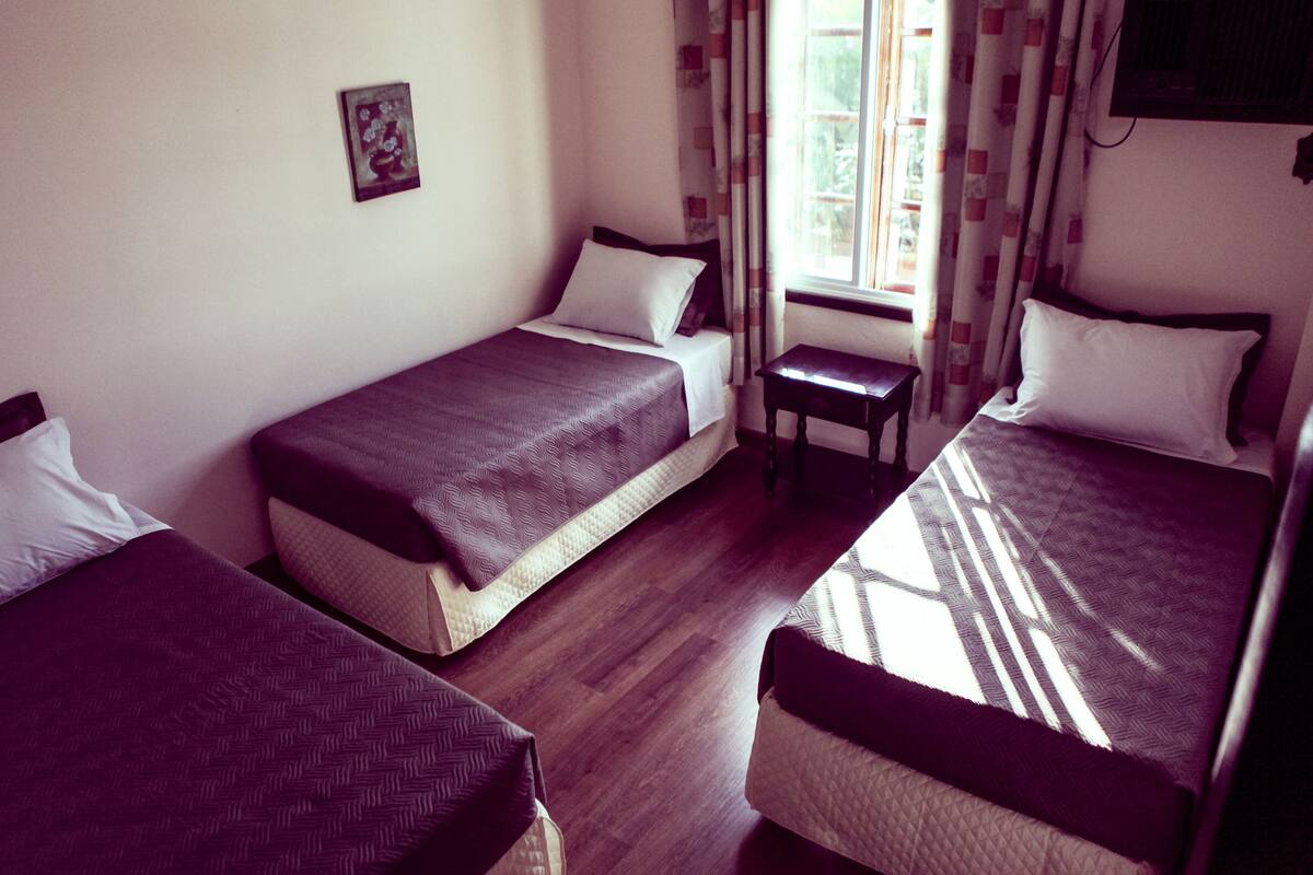 A room with three beds, each with a different patterned bedspread, a small table, and a window with curtains.