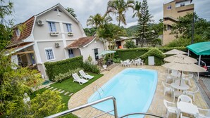 A swimming pool with lounge chairs and umbrellas, surrounded by greenery and a building with a sloped roof. - Hotel Blumenhof (Blumenau)
