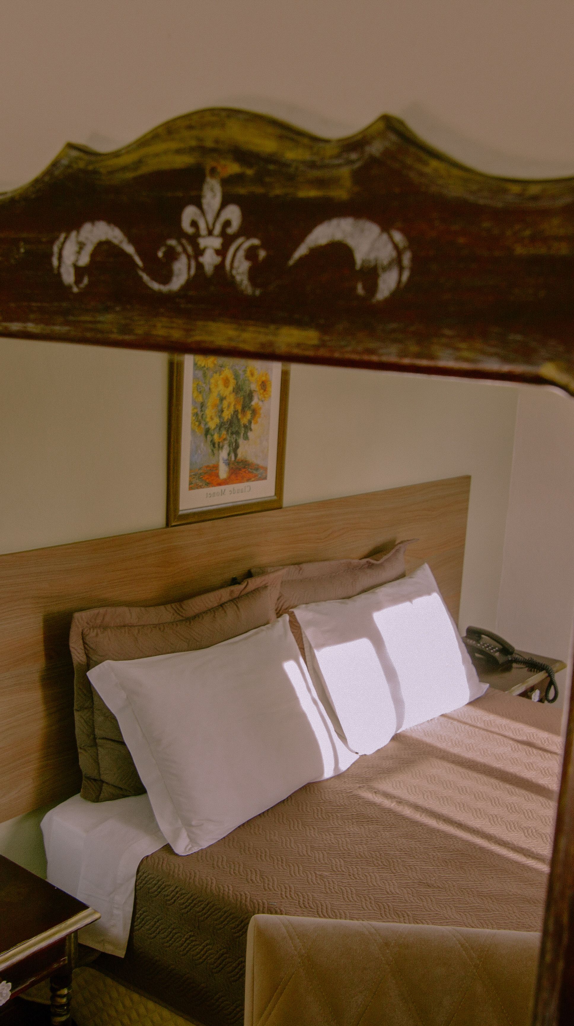 A neatly made bed with a wooden headboard, a bedside table with a telephone, and a framed painting on the wall.