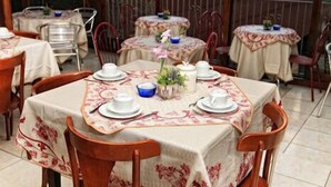 An outdoor dining area with tables set for two, covered with floral tablecloths, and surrounded by wooden chairs. - Hotel Blumenhof (Blumenau)