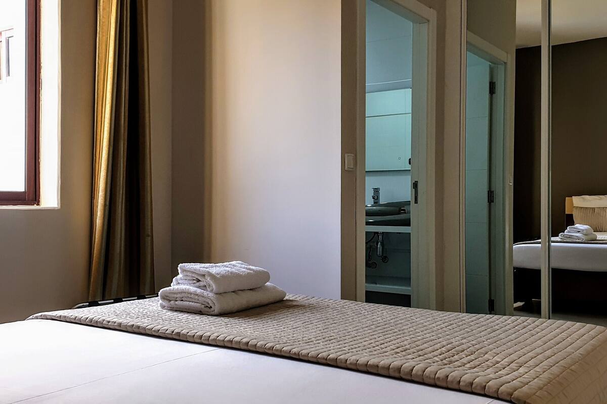 Ein Hotelzimmer mit Bett, einem Fenster mit Vorhängen, einem durchgegangenen Tür angedeuteten Badezimmer und einem Handtuch auf dem Bett.