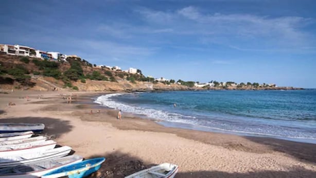 Una playa con barcos alineados en la arena, cielo despejado y edificios en una colina al fondo.