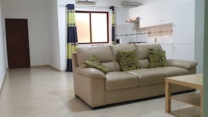 Una sala de estar con un sofá color beige, una mesa de centro de madera y una zona de cocina con armarios blancos y un microondas.
