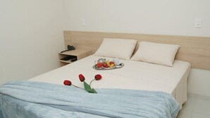 Una cama bien hecha con un cabecero de color claro, una mesita de noche con una lámpara, y una bandeja con fruta y una taza sobre la cama.