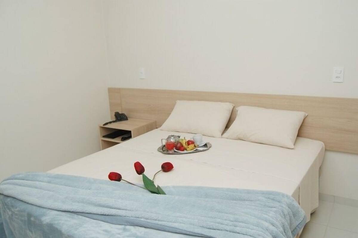 Een net opgemaakt bed met een lichtkleurig hoofdeinde, een nachtkastje met een lamp, en een schaal fruit en een kopje op het bed.