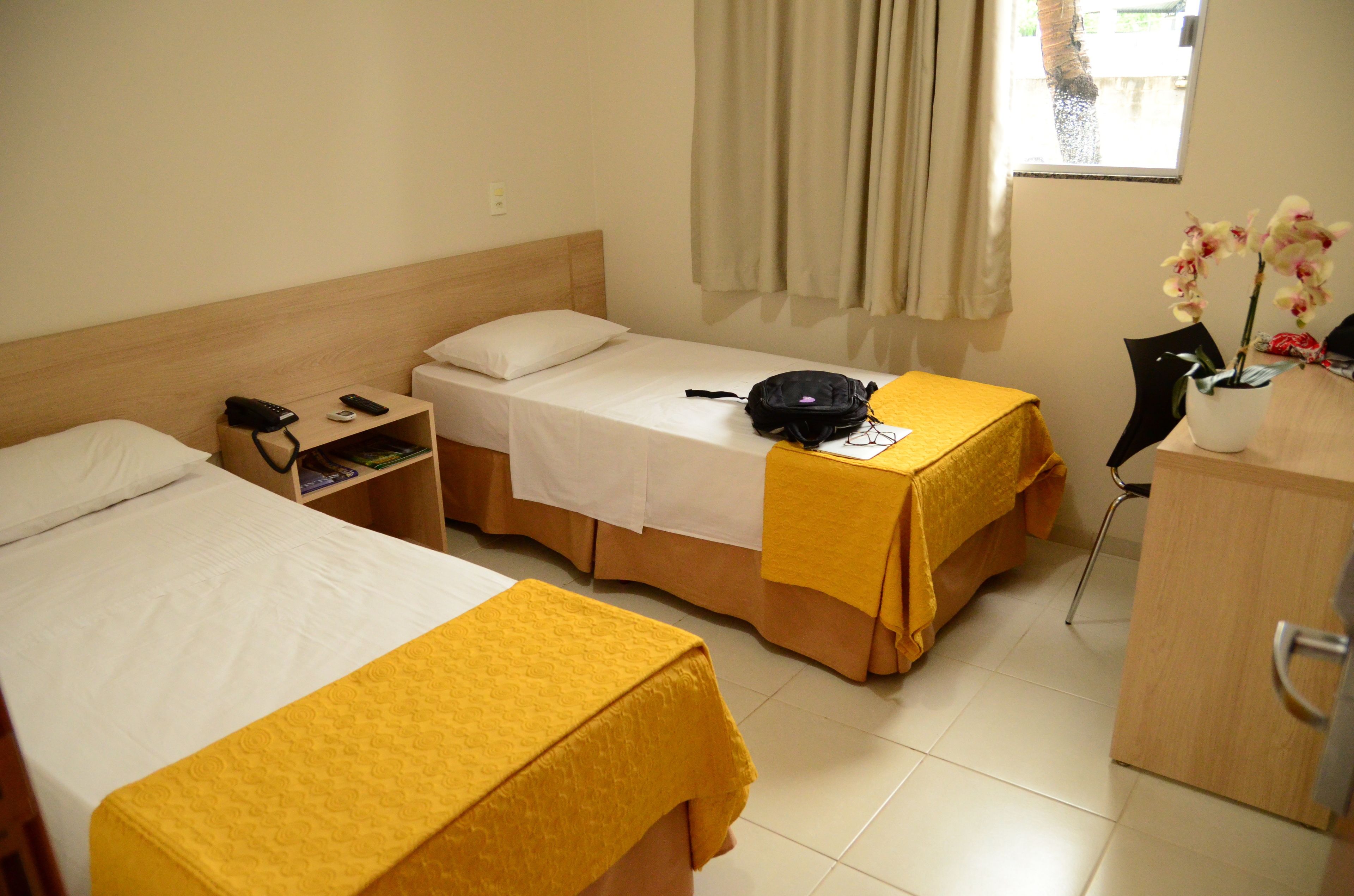 Ein Hotelzimmer mit zwei Einzelbetten, einem Nachttisch mit Lampe, einem kleinen Schreibtisch mit Stuhl und einem Fenster mit Vorhängen.