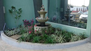 Una fuente de varios niveles en un jardín ajardinado con un muro verde y un ventanal que refleja un coche y edificios.