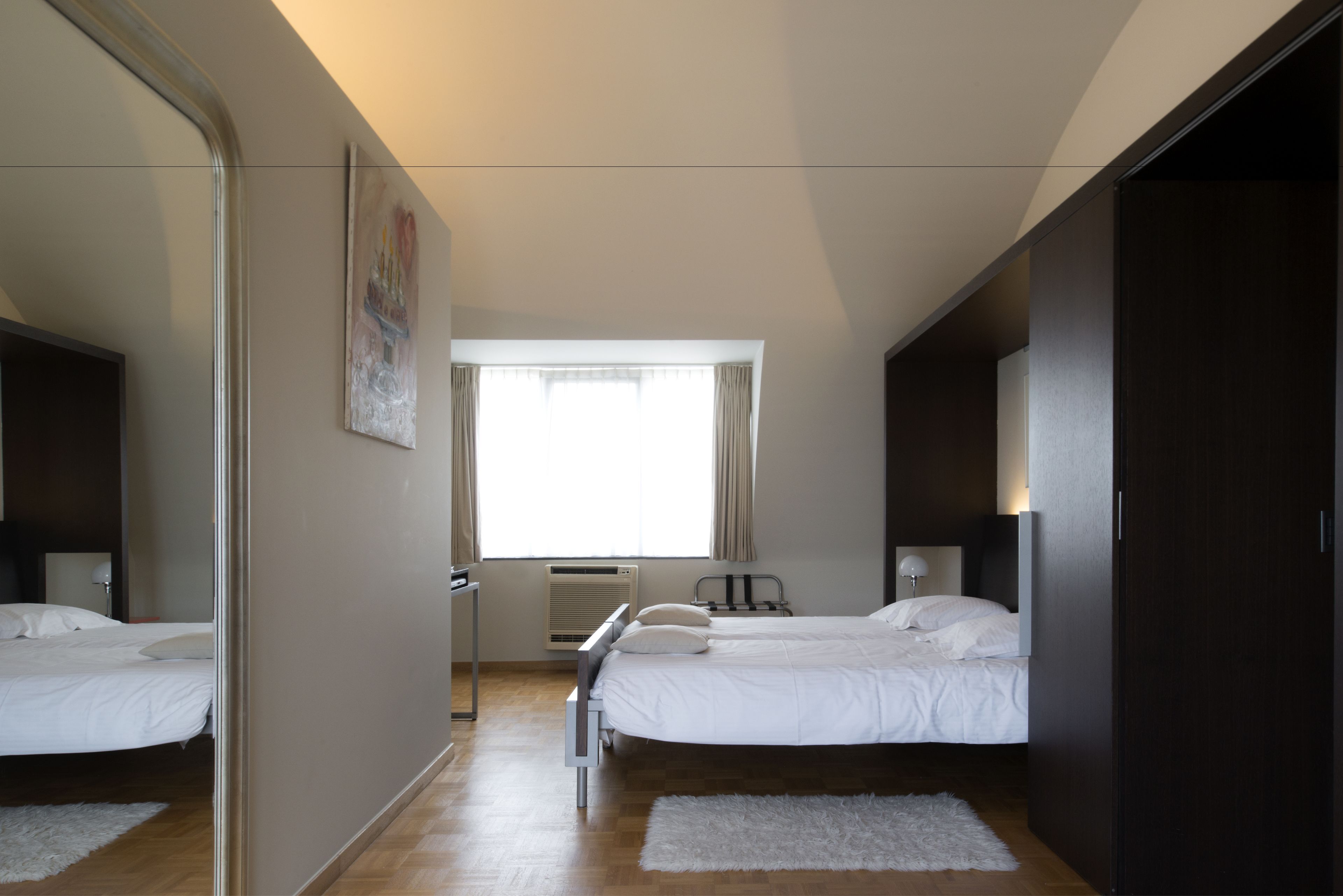 Un dormitorio con dos camas, un ventanal con cortinas, un radiador y una alfombra blanca.