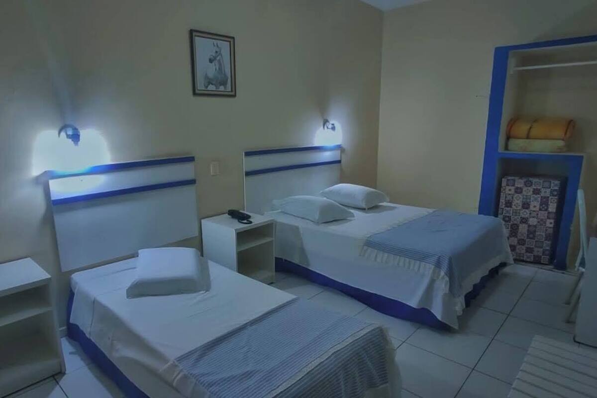 A hotel room with two beds, a nightstand, a desk, a wardrobe, and a small shelf with a lamp.