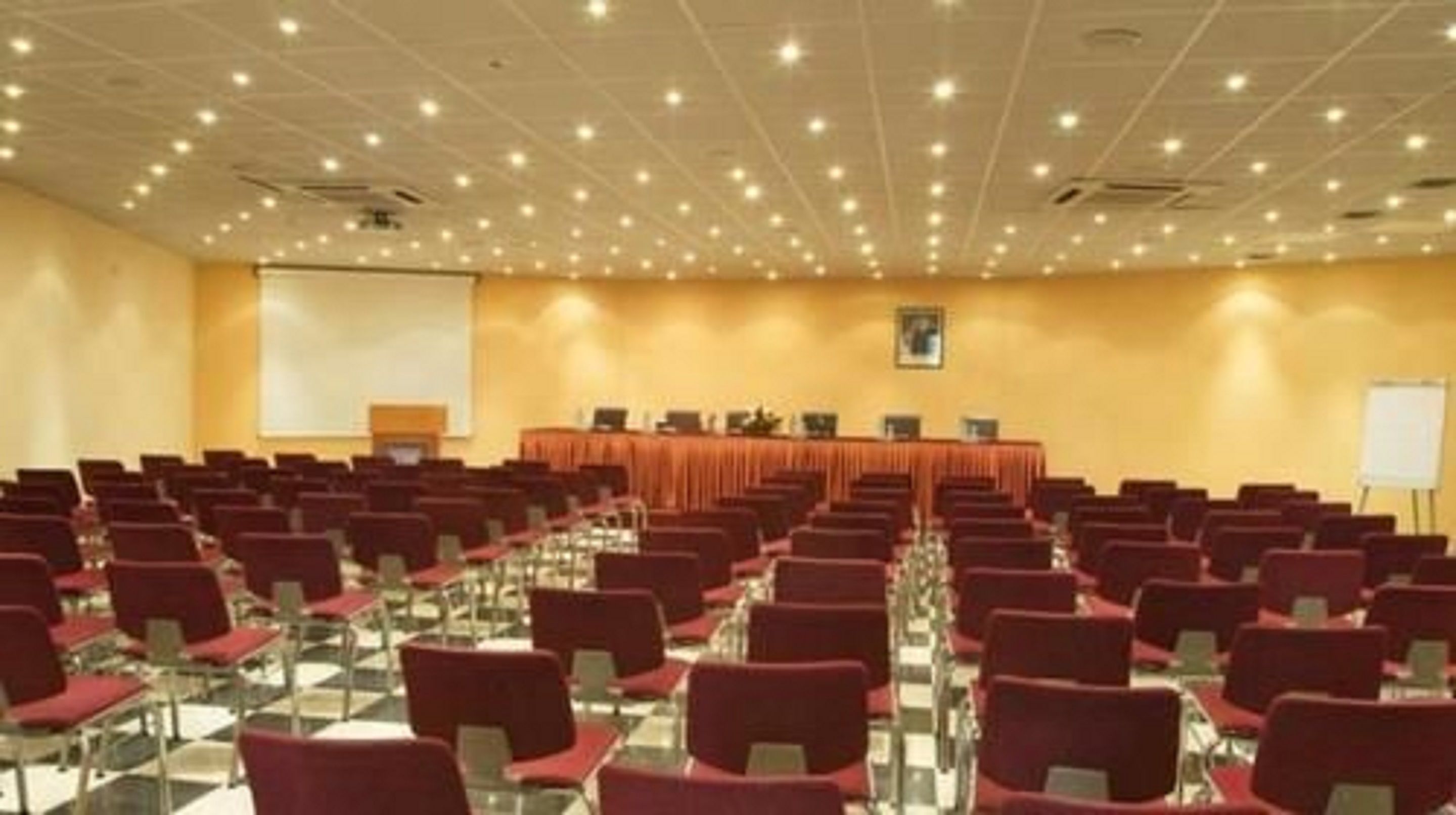A conference room with rows of red chairs, a long table at the front, and a projector screen.
