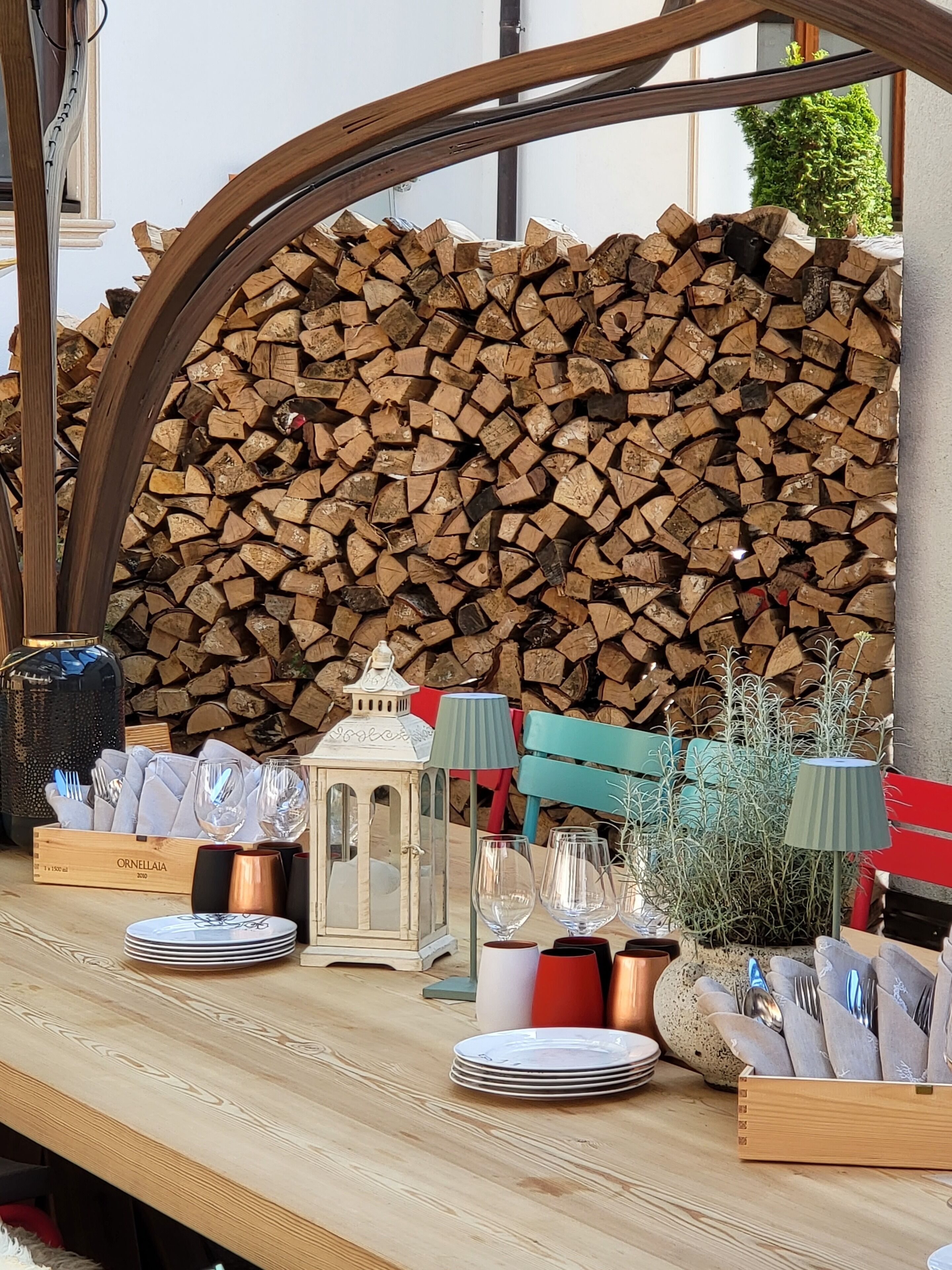 Una mesa de madera dispuesta con platos, cubiertos y un farol, con un fondo de leña apilada y una planta verde.