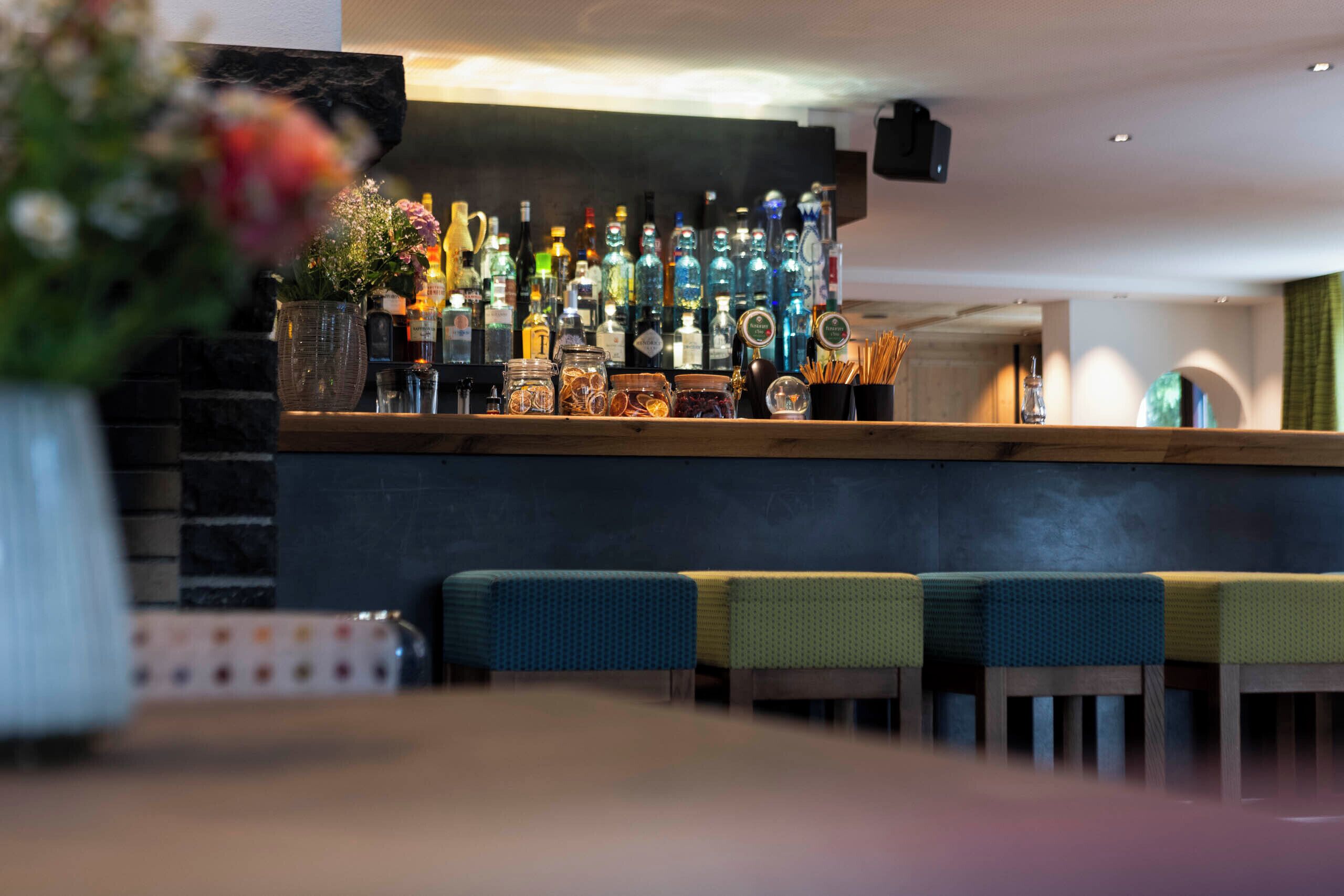 A bar area with a variety of bottles and glasses, a vase with flowers, and a seating area with cushioned chairs.