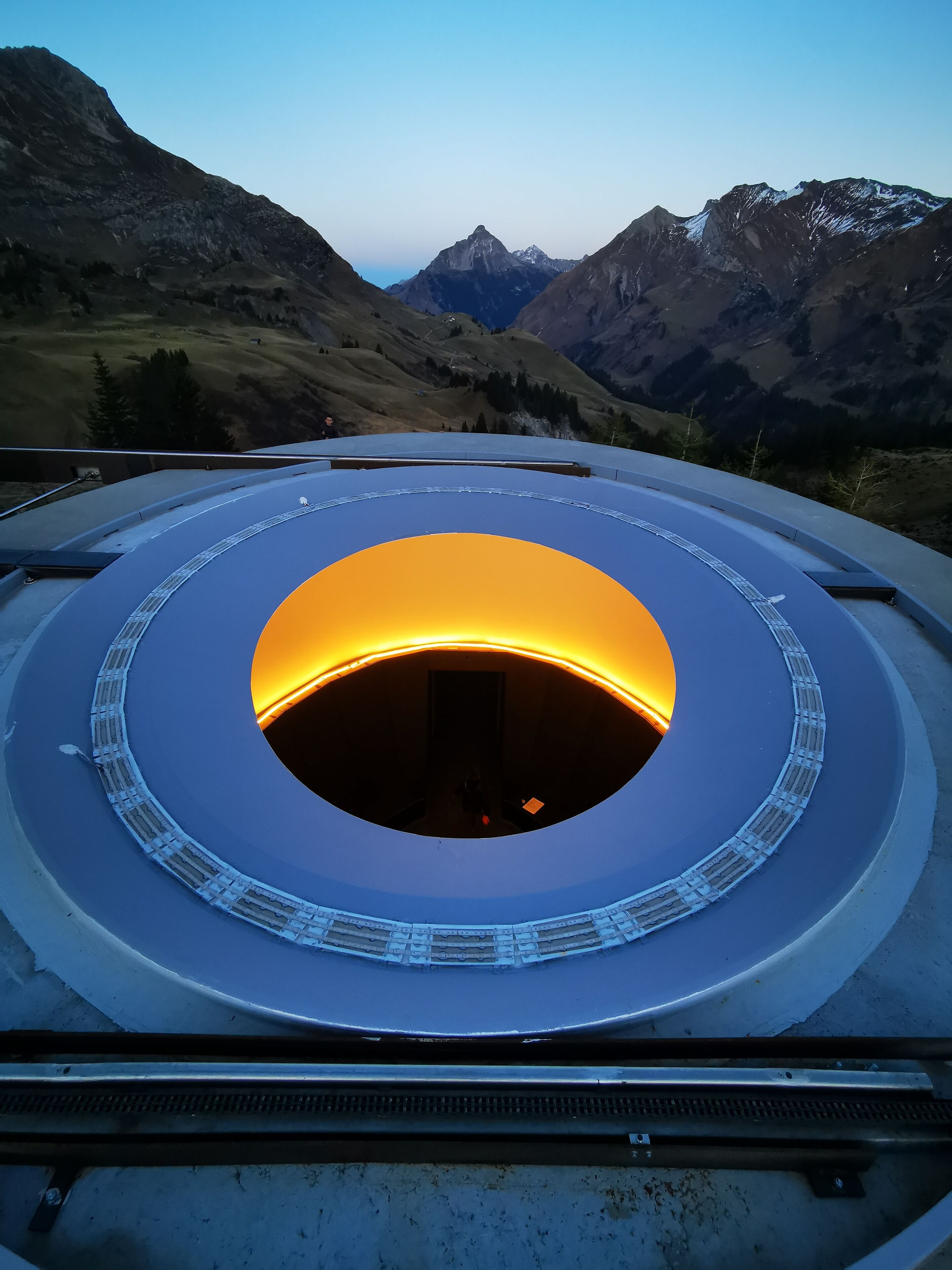 A circular structure with a central opening, surrounded by a railing, set against a mountainous backdrop.
