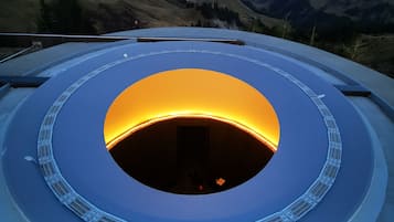 Una estructura circular con una abertura central, rodeada por una barandilla, con montañas al fondo.