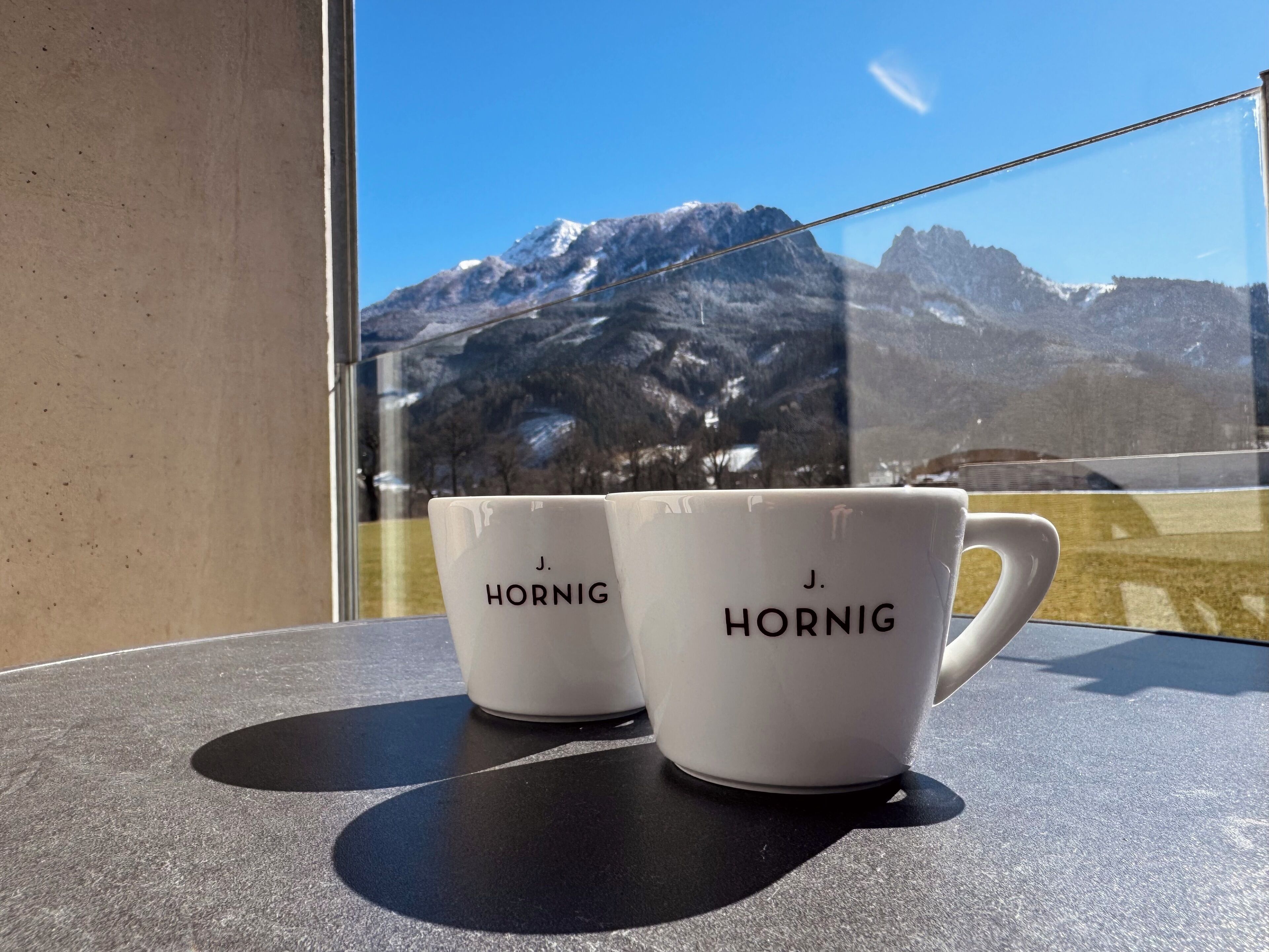 Zwei weiße Tassen mit dem Aufdruck 'J. HORNIG', platziert auf einer spiegelnden Oberfläche mit Bergblick im Hintergrund.