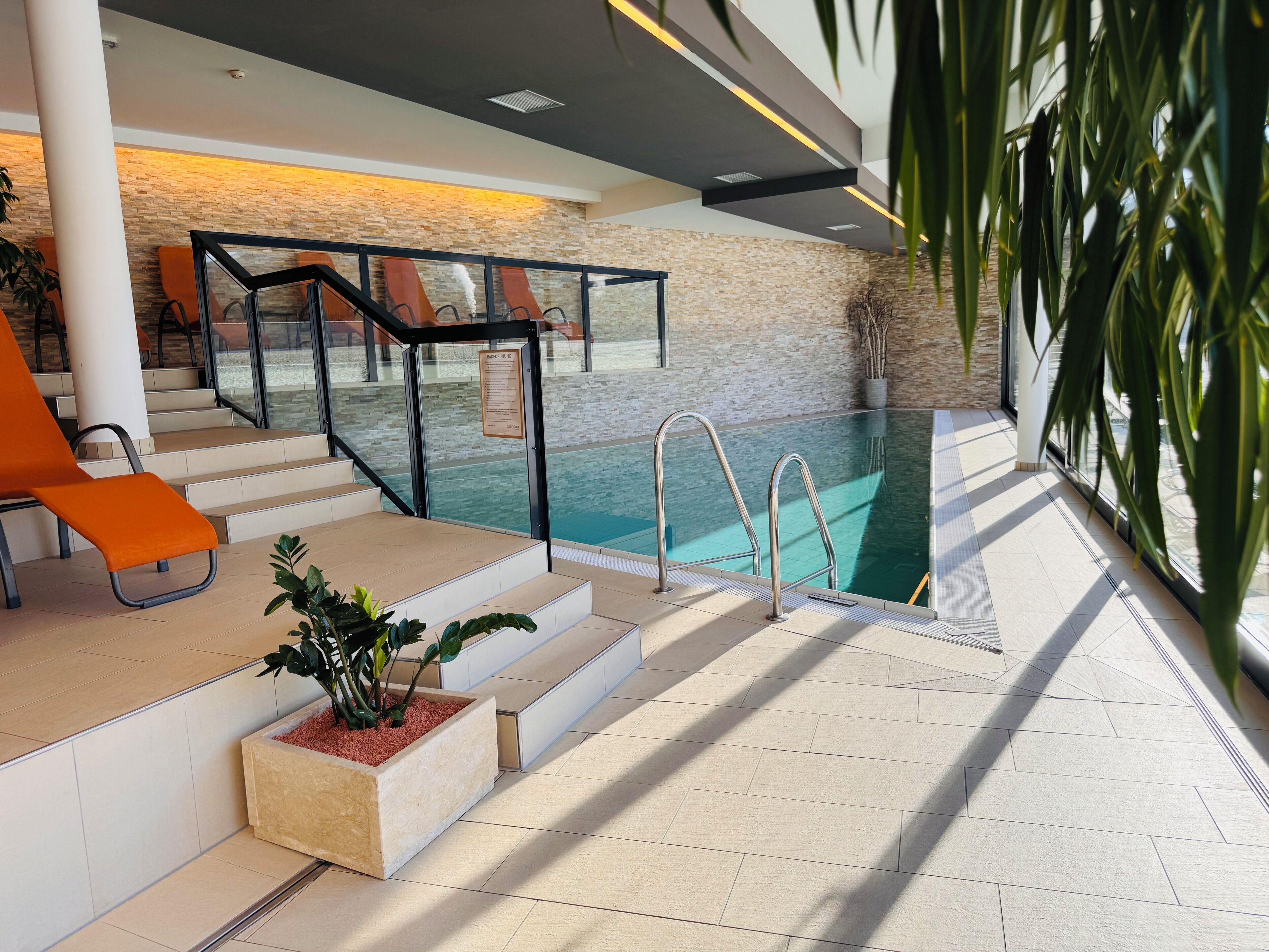 An indoor swimming pool with a glass barrier, steps leading to the pool, and a seating area with orange chairs.