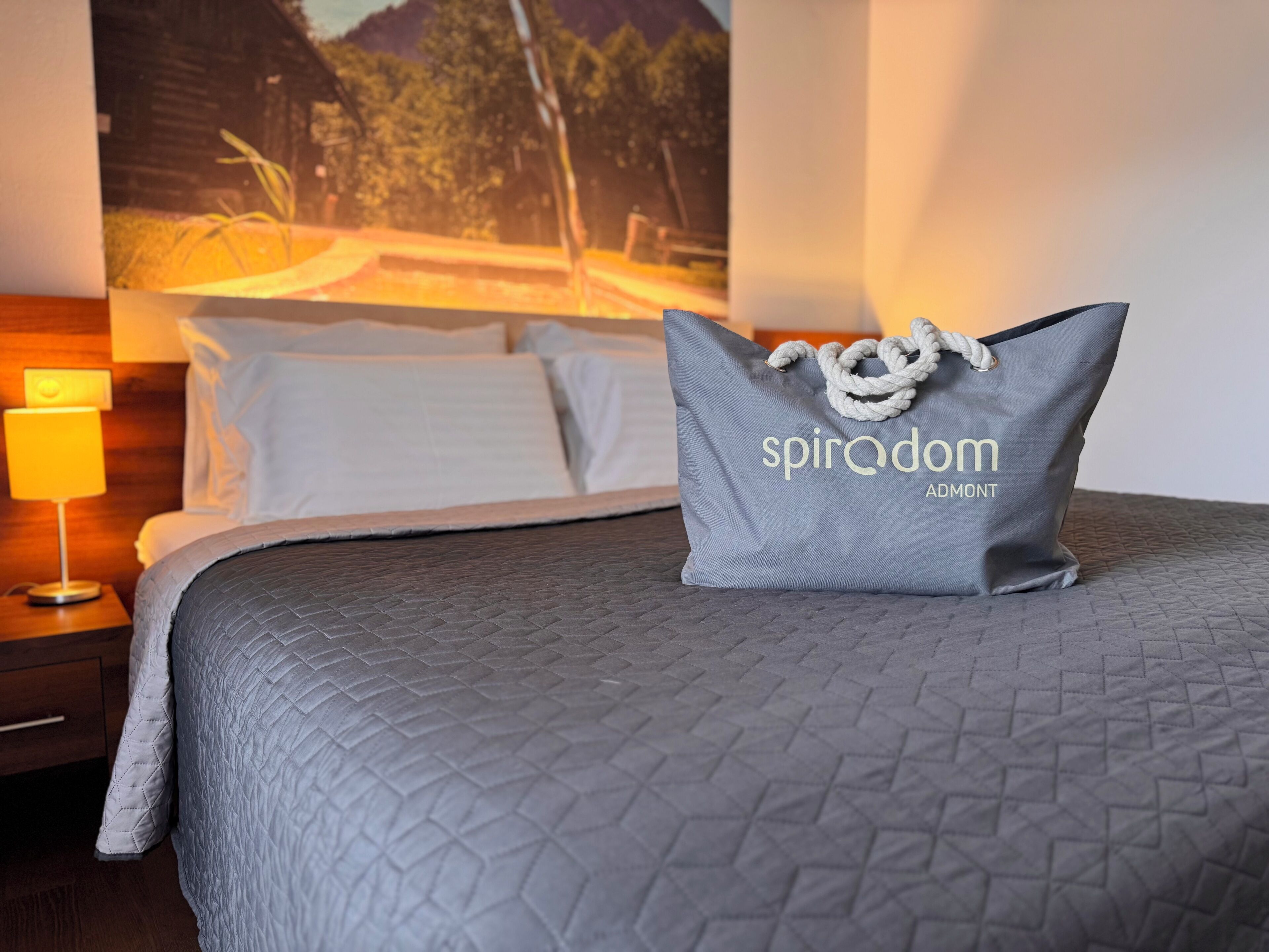 A neatly made bed with a grey quilt, a pillow with the text "spiradom ADOMONT", and a nightstand with a lamp.