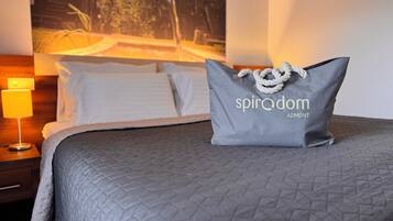 A neatly made bed with a grey quilt, a pillow with the text "spiradom ADOMONT", and a nightstand with a lamp.