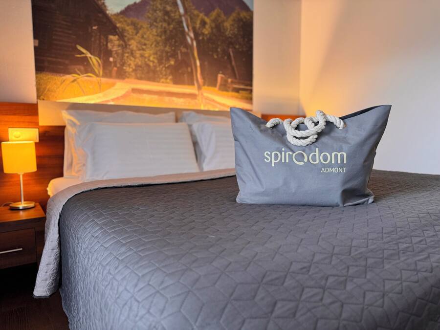 A neatly made bed with a grey quilt, a pillow with the text "spiradom ADOMONT", and a nightstand with a lamp.