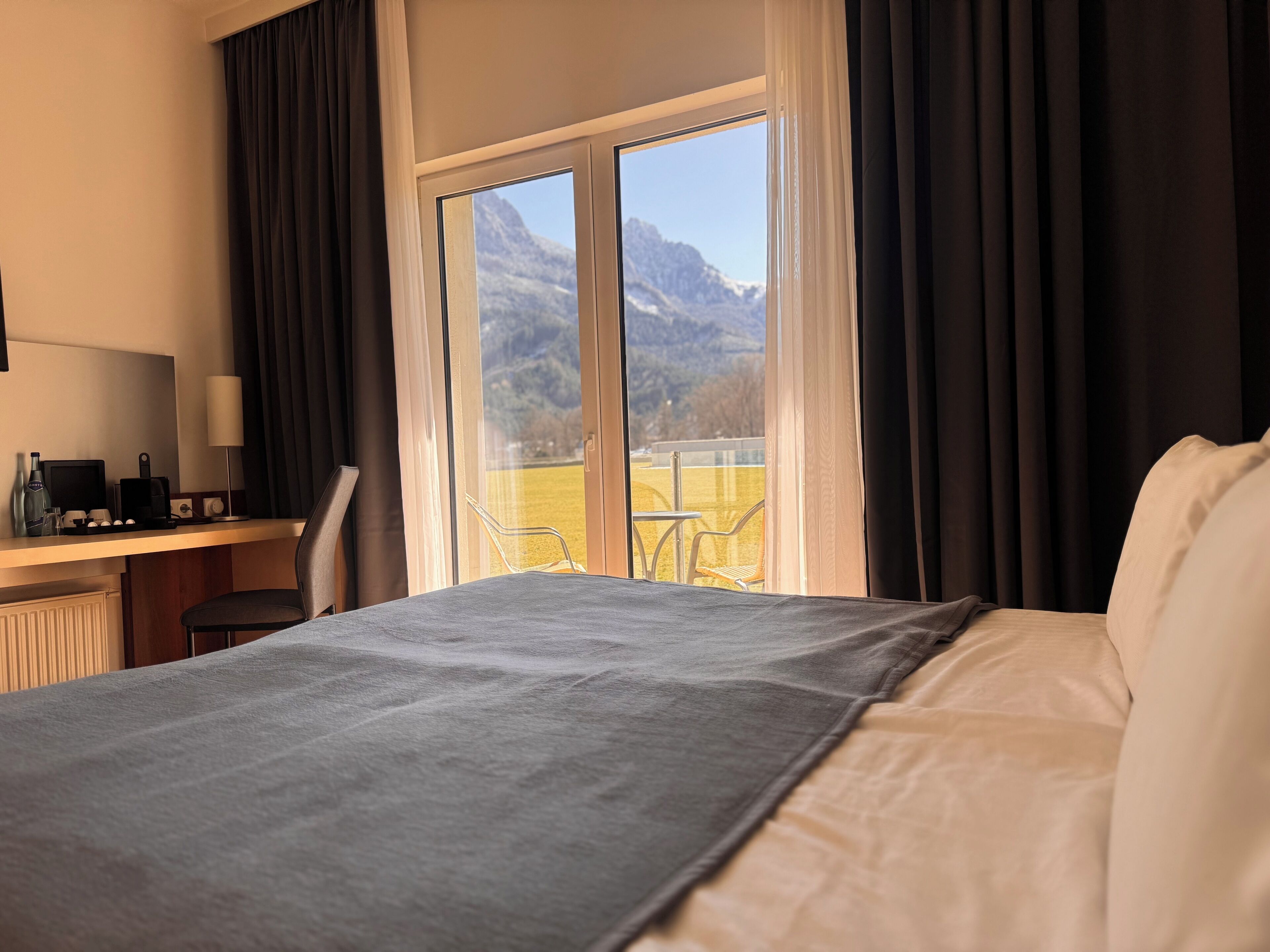 Ein Hotelzimmer mit einem Bett, einem Schreibtisch mit Computer und Blick auf Berge durch große Fenster.