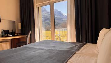 Habitación de hotel con una cama, un escritorio con ordenador y vistas a montañas a través de amplios ventanales.