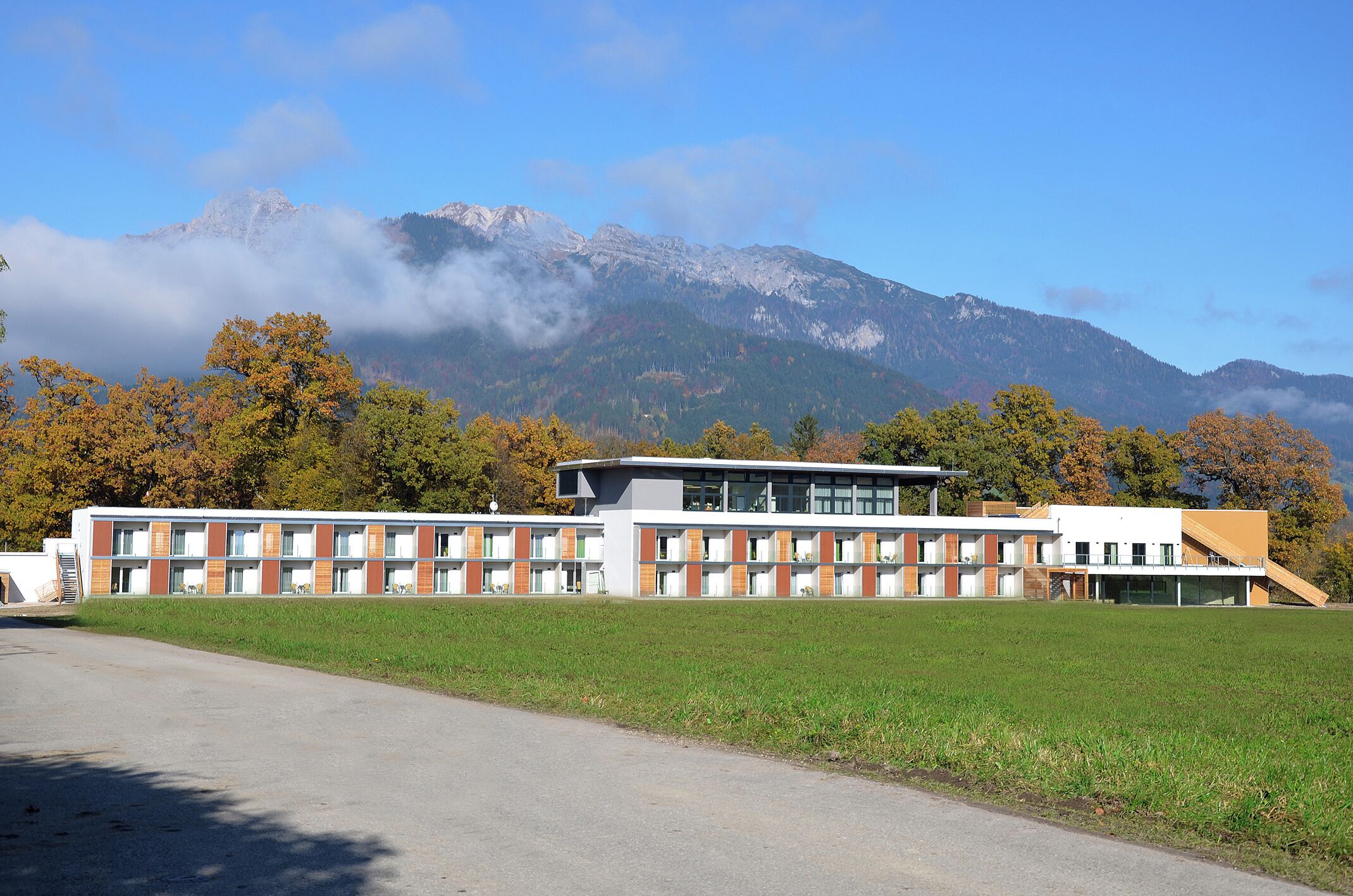 A modern building with a flat roof and large windows, surrounded by a grassy field and trees with autumn foliage.