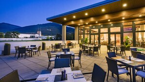 An outdoor dining area with tables and chairs, a covered section with large windows, and a view of a church and mountains in the background. - Hotel Spirodom Admont (Admont)