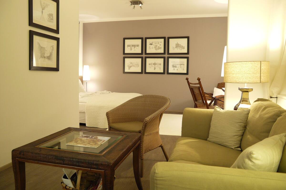 Ein Hotelzimmer mit einem Bett, einem grünen Sofa, einem Korbstuhl, einem Tisch mit Glasplatte und gerahmten Bildern an der Wand.