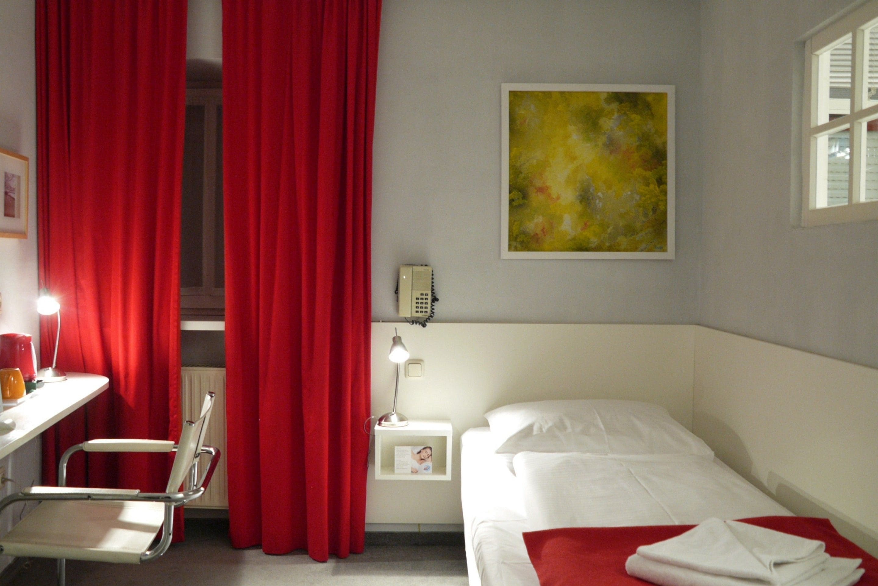 A hotel room with a bed, a desk with a chair, a nightstand with a lamp, and red curtains.