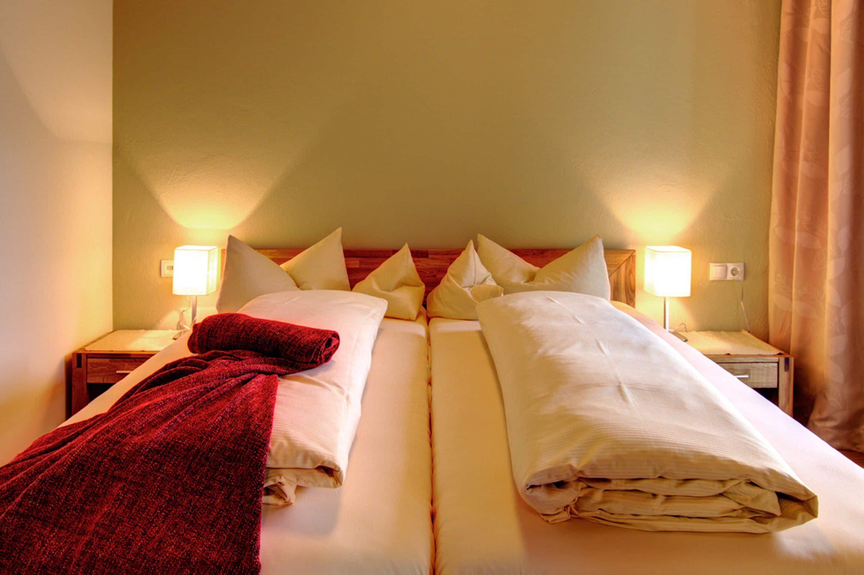 Una cama doble con una manta roja y ropa de cama blanca, dos lámparas de noche y un cabecero de madera.