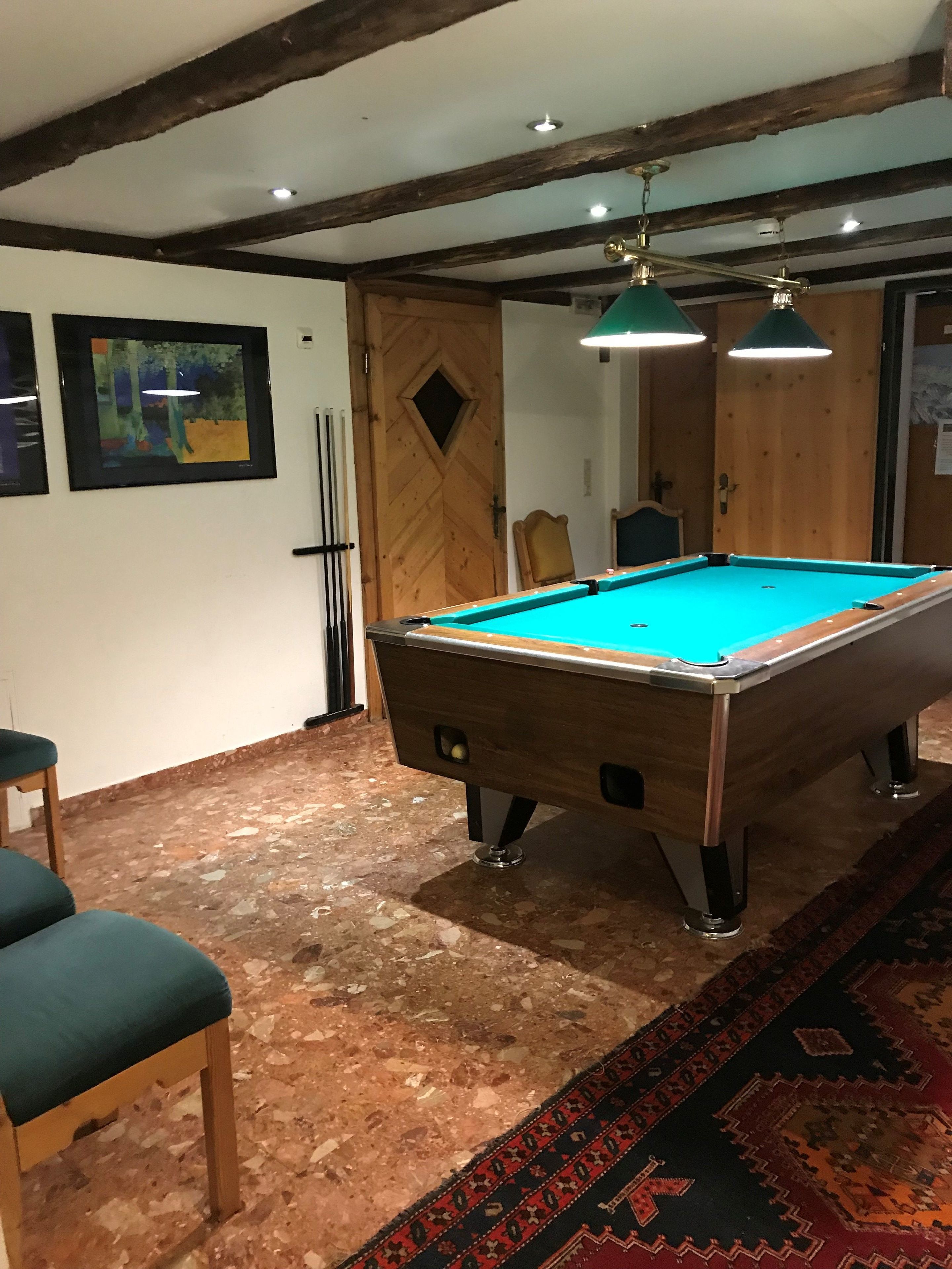 A pool table with a green felt surface, wooden legs, and a light fixture above it. There are two pool cues leaning against the wall and a chair with a cushion in front of the table.
