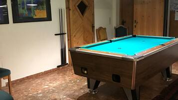 A pool table with a green felt surface, wooden legs, and a light fixture above it. There are two pool cues leaning against the wall and a chair with a cushion in front of the table.
