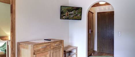 A bedroom with a wooden dresser, a bed with a green blanket, a mounted TV, and a wooden door.