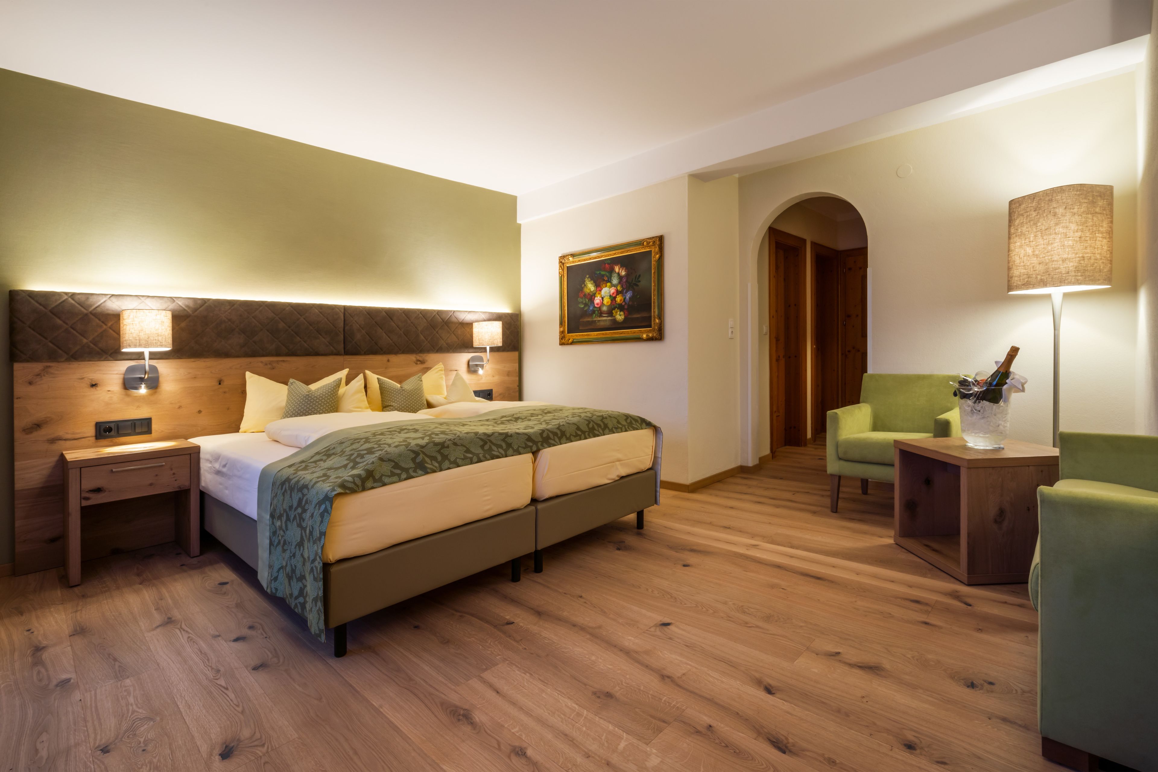 Une chambre d’hôtel avec un lit, des tables de chevet, un fauteuil vert, une table d’appoint et un tableau accroché au mur.