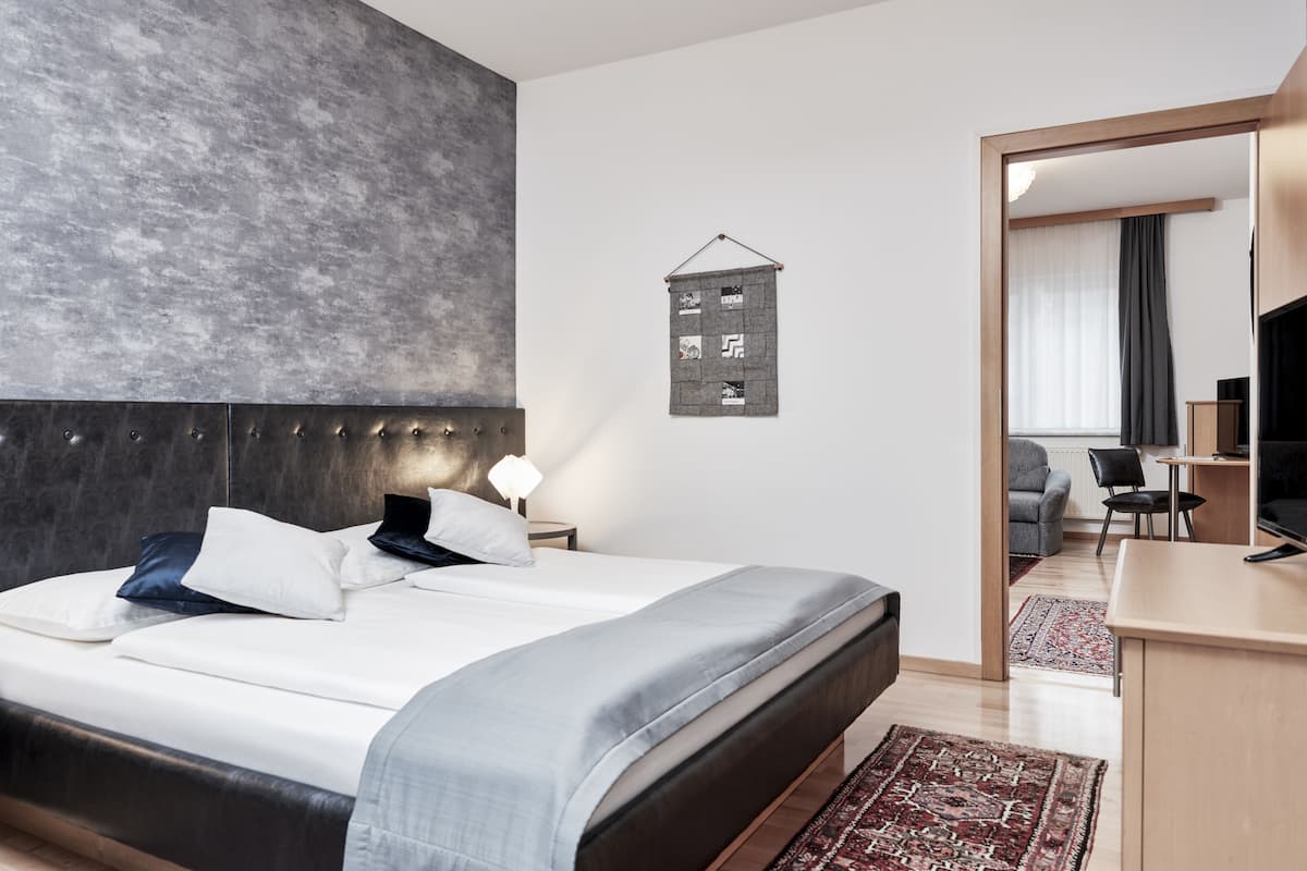 A modern hotel room with a large bed, a headboard, a small table with a lamp, a TV, and a view into the living area.