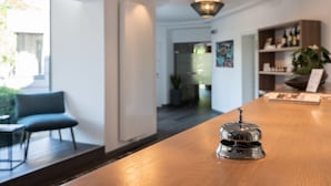 A hotel reception desk with a wooden surface, a silver bell, and a view of a modern interior including a chair and a plant. - Hotel Garni Birkenhof (Bad Radkersburg)