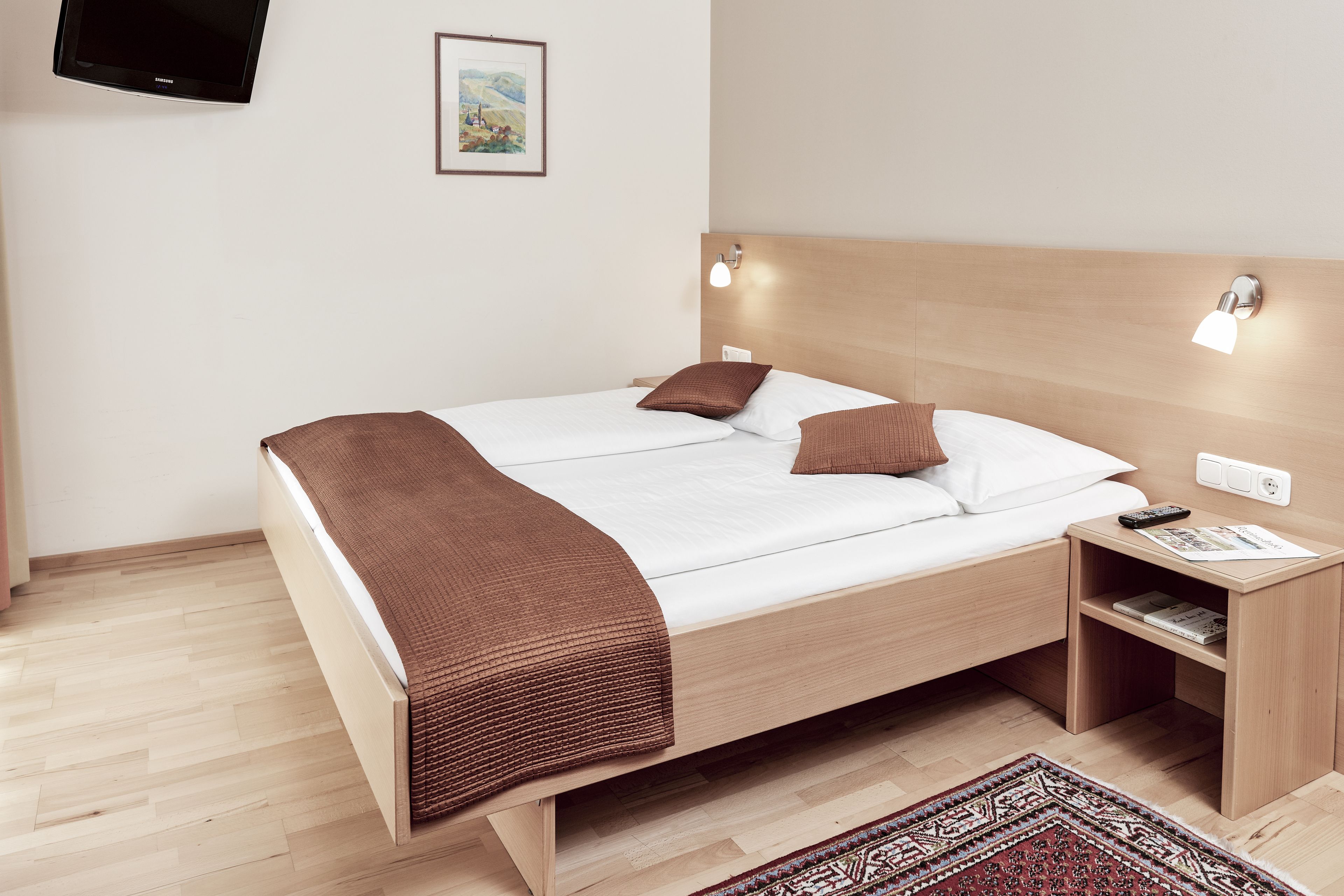 Ein modernes Hotelzimmer mit einem hölzernen Bett, zwei Nachttischlampen, einem Flachbildfernseher und einem kleinen Nachttisch mit Fernbedienung.