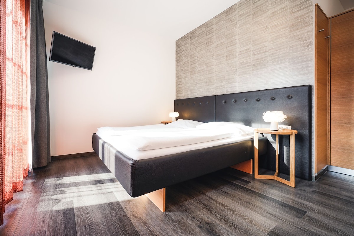 A modern hotel room with a bed, a bedside table with a lamp, a flat-screen TV mounted on the wall, and a wooden wardrobe.