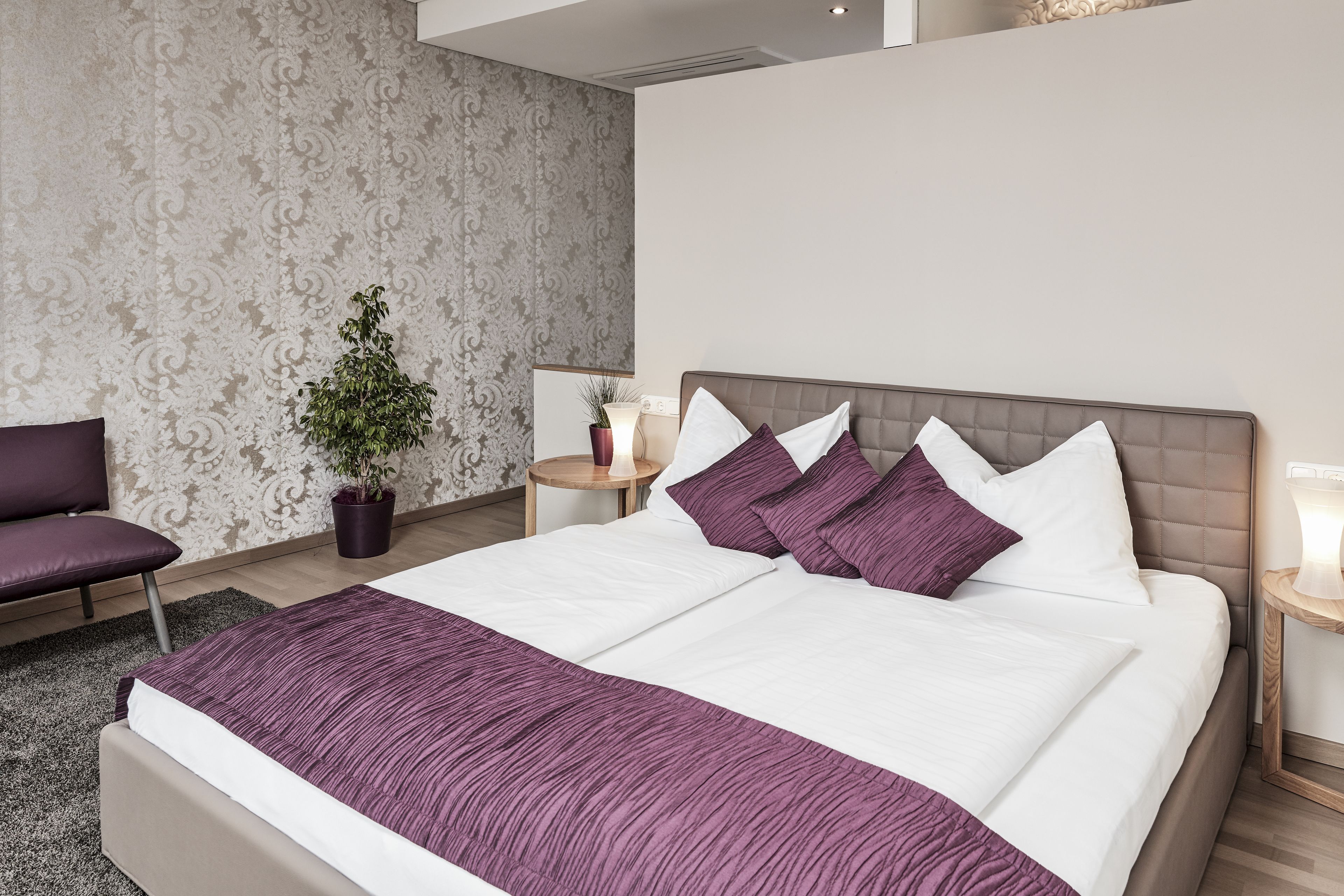 Una habitación de hotel con una cama grande, una mesita de noche, una silla, una planta en maceta y un papel pintado con motivos.