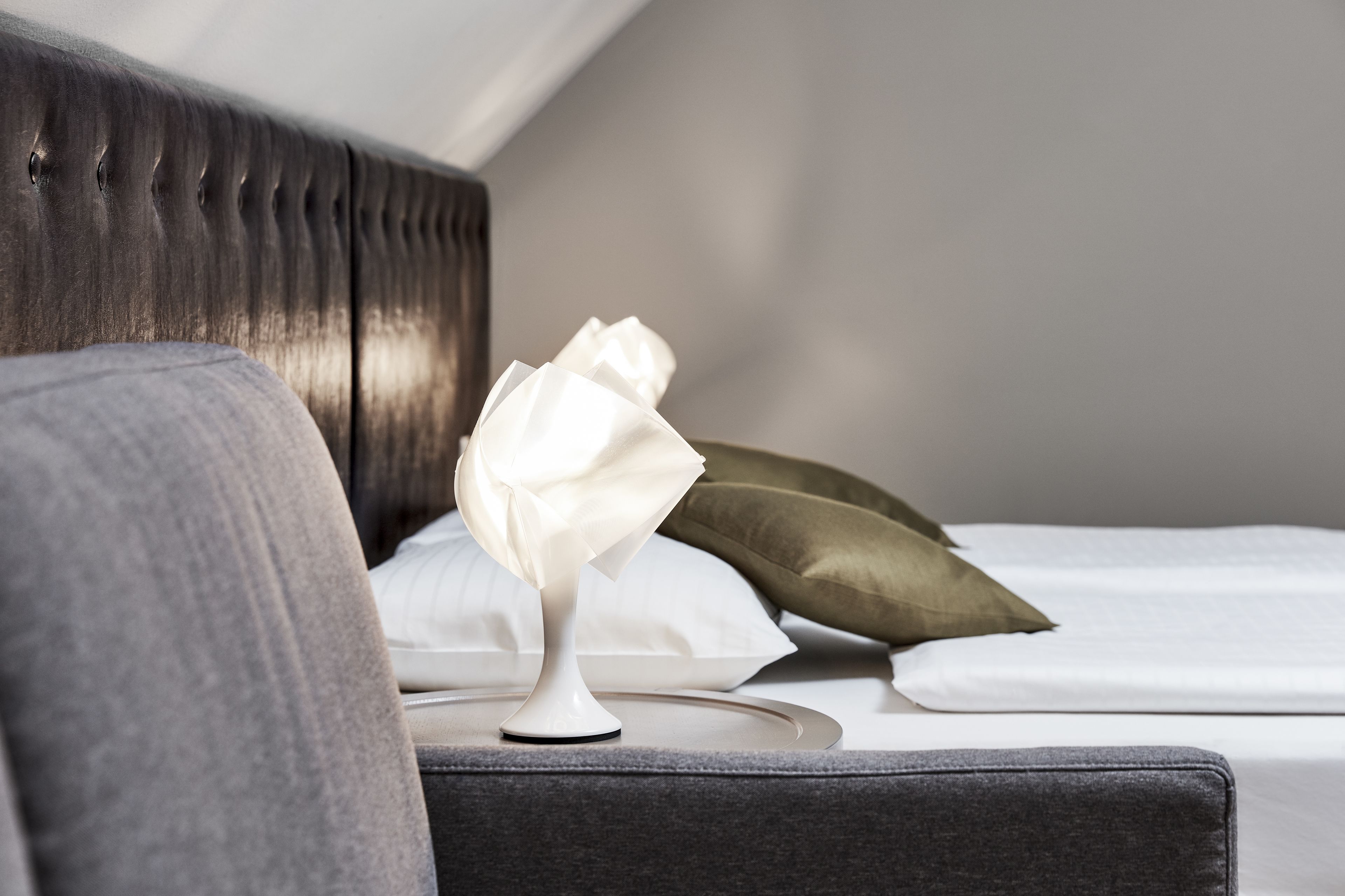 Una cama bien hecha con sábanas blancas, un cabecero tapizado en gris y una mesita de noche blanca con una lámpara blanca.
