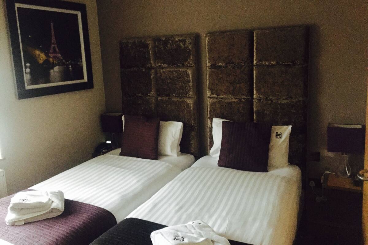 A hotel room with two beds, a stone headboard, a framed picture of the Eiffel Tower, and a nightstand with a lamp.