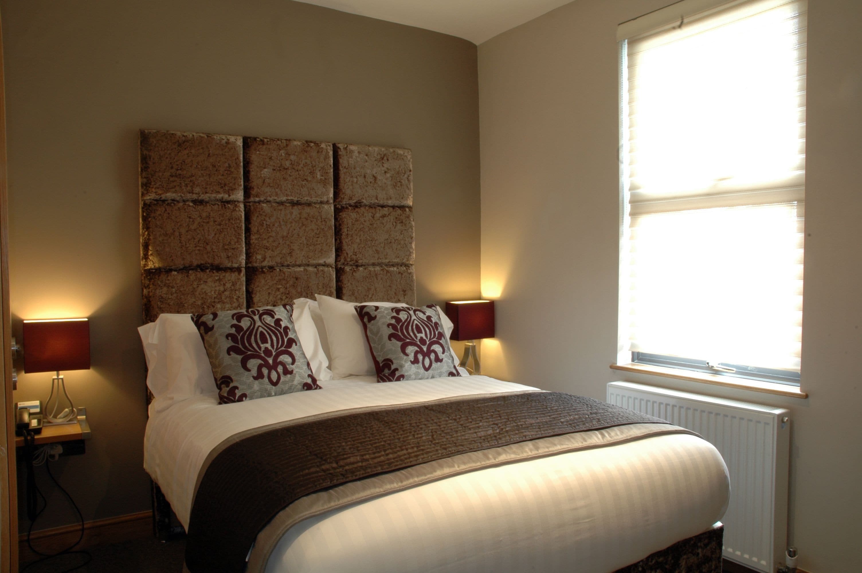 A neatly made bed with a headboard, two bedside lamps, and a window with blinds.