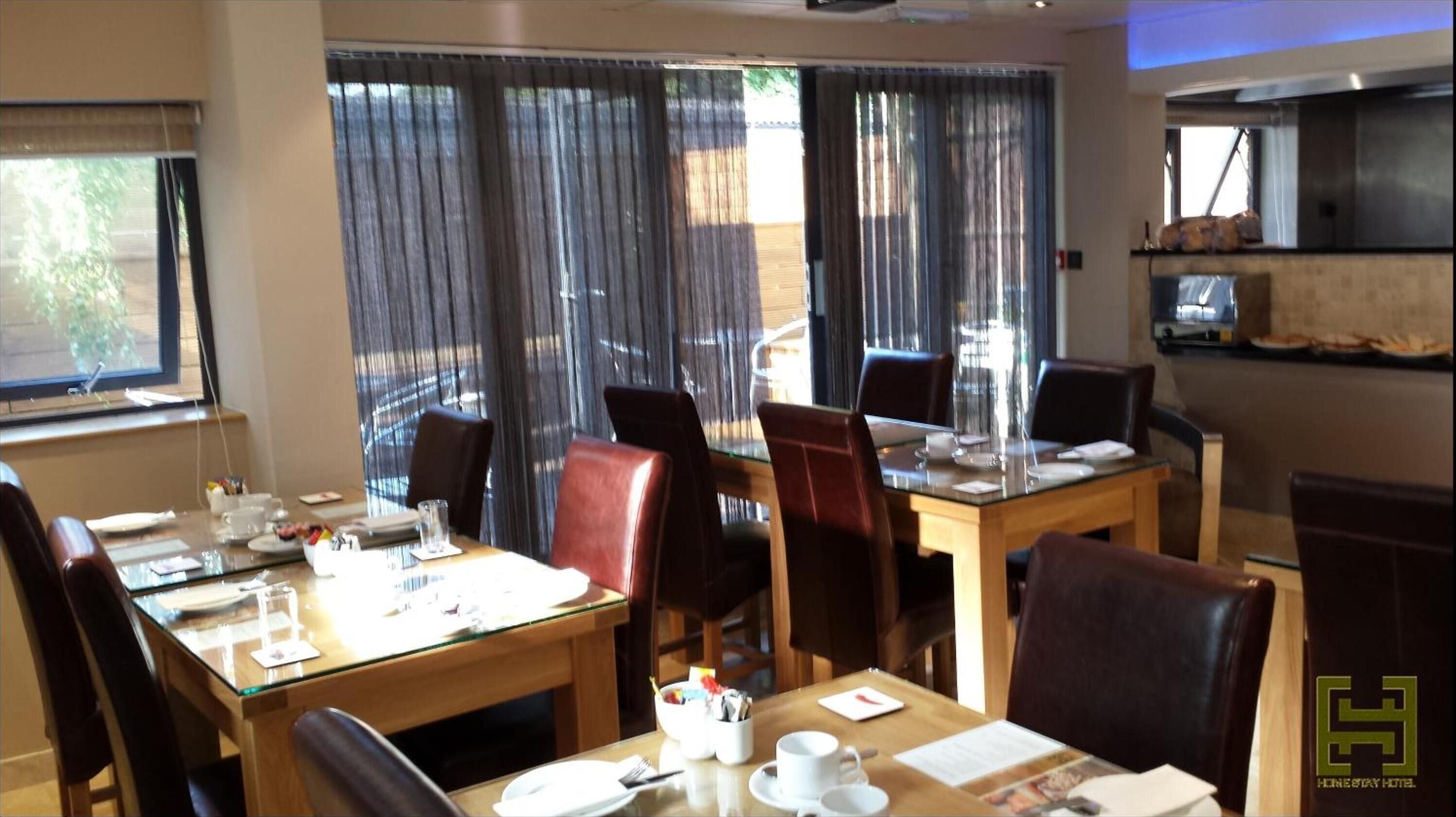 A hotel dining area with a glass table, leather chairs, and a view through large windows with curtains.