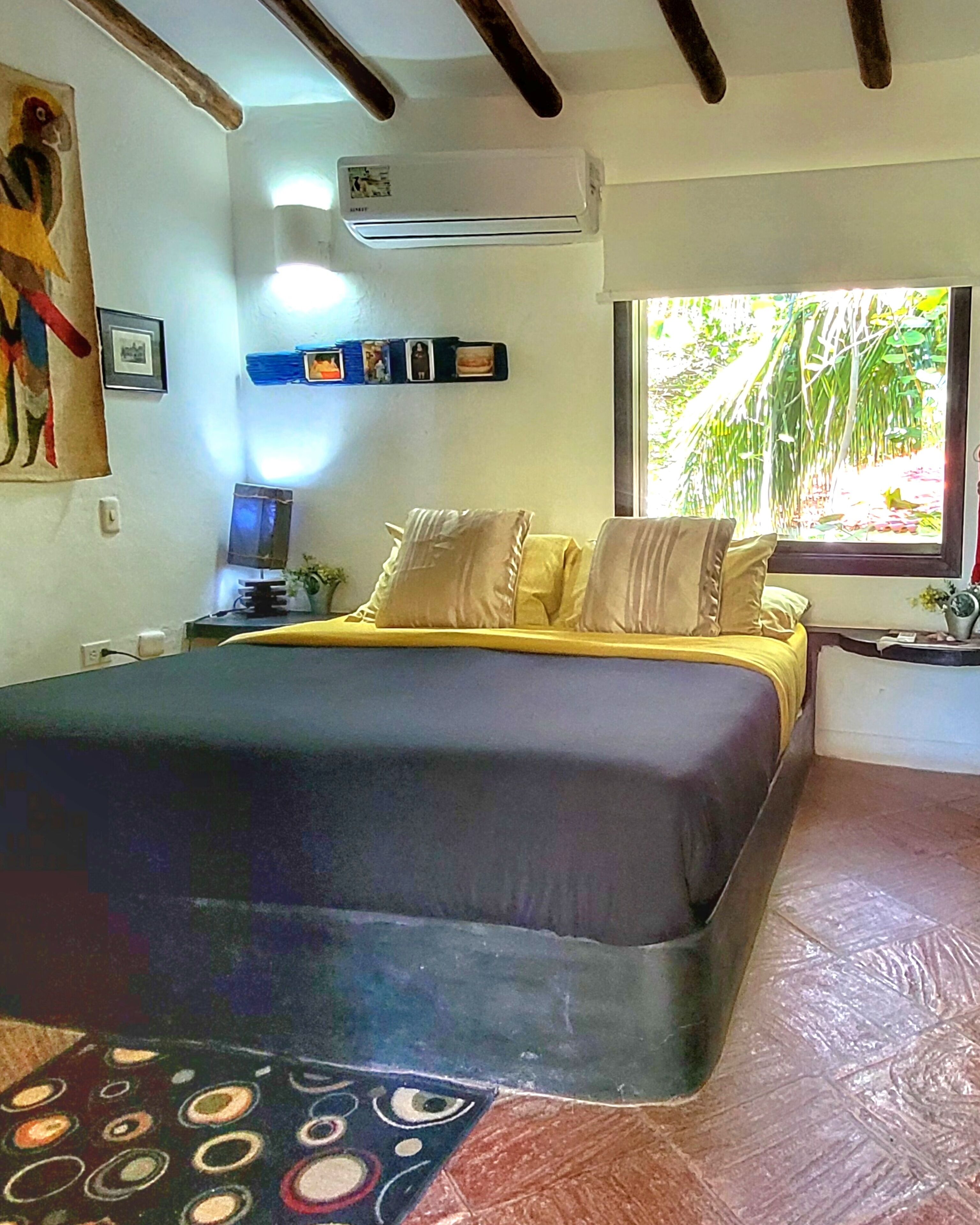 A bedroom with a bed, a nightstand, a window with a view of greenery, and a wall-mounted air conditioner.