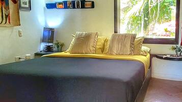 A bedroom with a bed, a nightstand, a window with a view of greenery, and a wall-mounted air conditioner.